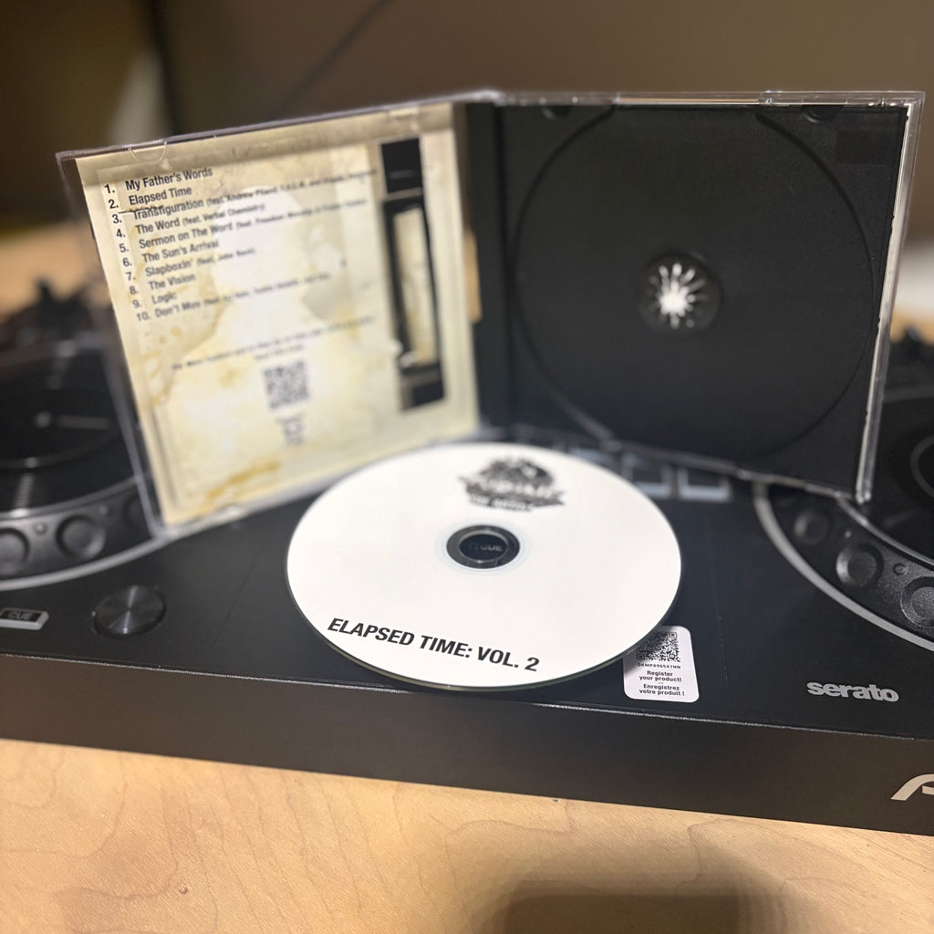 A CD case opened to show a printed tracklist insert, a black CD disc, and a white CD disc labeled 'ELAPSED TIME: VOL. 2' resting on a black Serato DJ controller on a wooden surface.