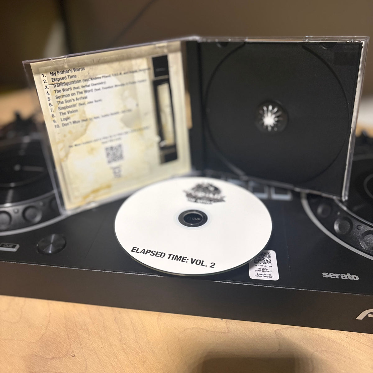 A CD case opened to show a printed tracklist insert, a black CD disc, and a white CD disc labeled 'ELAPSED TIME: VOL. 2' resting on a black Serato DJ controller on a wooden surface.